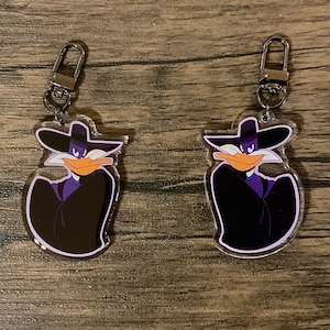 Könnte beinhalten: Zwei transparente Acryl-Schlüsselanhänger mit einem Cartoon-Enten-Motiv, das einen violetten Anzug und einen schwarzen Hut trägt. Die Ente hat einen schelmischen Ausdruck und hält eine Pistole in der Hand. Die Schlüsselanhänger sind an silbernen Metallschlüsselringen befestigt.