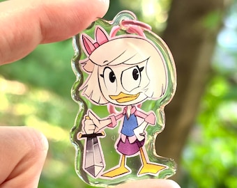 May include: A clear acrylic keychain featuring a cartoon character with blonde hair, a pink bow, and a blue shirt holding a silver sword. The character is standing on a clear acrylic base.