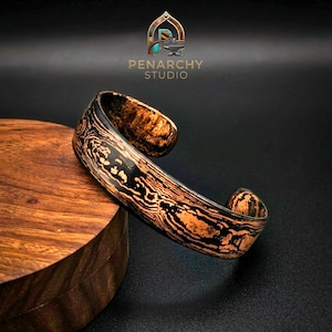 Puede incluir: Una pulsera tipo brazalete con un patrón de vetas de madera en capas en tonos contrastantes de negro y cobre. La superficie metálica muestra una estética orgánica y texturizada con un acabado pulido. El logotipo de Penarchy Studio es visible.