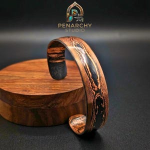 May include: A cuff bracelet featuring a layered metal pattern with alternating copper-toned and dark black bands. The surface displays a wood grain-like texture created by the metal layers. The logo PENARCHY STUDIO is visible at the top.