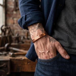 Pode incluir: Uma pulseira de cobre martelado com acabamento polido usada no pulso. A pulseira apresenta uma superfície texturizada e irregular e um design simples aberto. O metal tem um tom metálico castanho-avermelhado quente que reflete a luz.