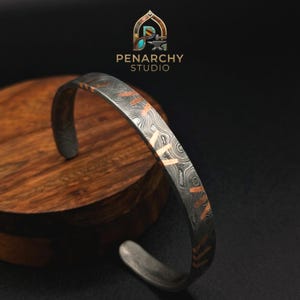May include: A metal cuff bracelet featuring a dark gray Damascus steel pattern with swirling lines and inlaid copper-colored accents. The bracelet has an open-ended design and displays the text PENARCHY STUDIO at the top of the image.