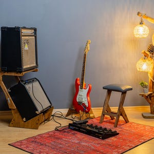 Può includere: Un'attrezzatura da studio musicale con una chitarra elettrica rossa, due amplificatori neri su un supporto in legno, uno sgabello in legno con seduta grigia e una lampada da terra decorativa con paralumi intrecciati. Sono presenti anche un tappeto rosso e un pedale di effetti nero.