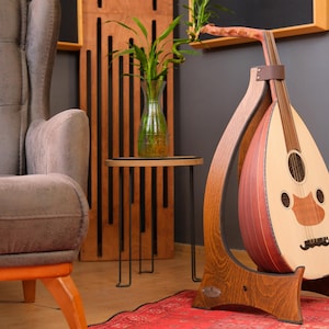 Puede incluir: Un oud de madera, un instrumento de cuerda de Oriente Medio, expuesto sobre un soporte de madera personalizado. El oud tiene una cara de color claro con una boca oscura marrón y una parte posterior marrón rojiza. El soporte es de madera oscura y tiene un diseño curvo.