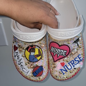 May include: A pair of white Crocs decorated with rhinestones and colorful patches. The patches include a nurse with a stethoscope, a heart with the word "NURSE" on it, and a red heart with a stethoscope. The words "NURSE" and "nursed" are also written on the shoes.