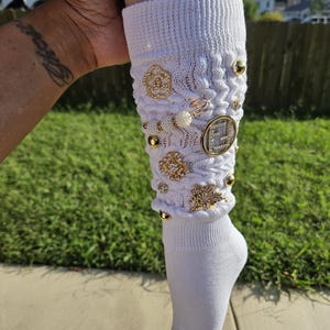 May include: White knee-high socks with a decorative design featuring gold and white embellishments. The embellishments include a large gold letter "E", small gold beads, and a gold flower.