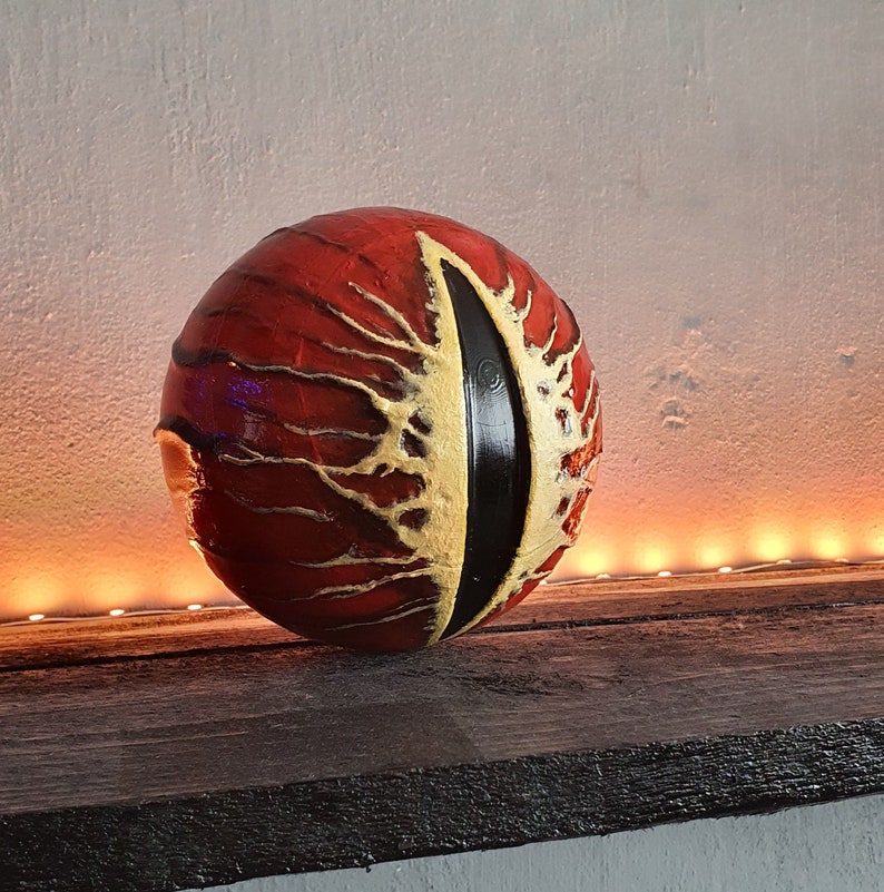 May include: A red and gold sphere with a black eye design. The sphere is sitting on a wooden surface.