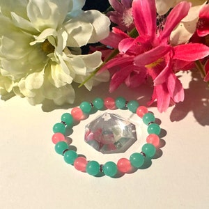 May include: A green and pink beaded bracelet with silver accents. The bracelet is lying on a white surface with a clear crystal in the background.
