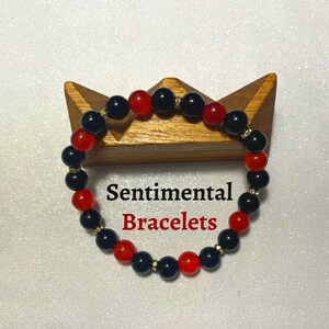 May include: A red and black beaded bracelet with gold accents. The bracelet is on a wooden stand. The text "Sentimental Bracelets" is visible below the bracelet.