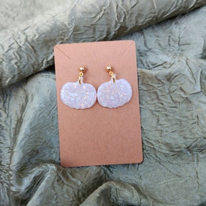 May include: A pair of white pumpkin-shaped earrings with iridescent glitter embedded in the surface. The earrings have gold-colored posts and are displayed on a brown card against a textured, olive-green background.