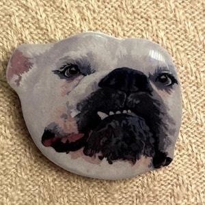 May include: A white and gray bulldog face design. The bulldog has a black nose and mouth, with a pink tongue. The eyes are dark and expressive. The background is a light beige.