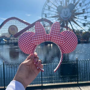 May include: A red and white gingham patterned headband with mouse ears and a bow. The ears and bow are made of a soft, padded fabric. The headband is white and has a curved shape.