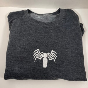May include: A dark gray t-shirt with a white embroidered Venom symbol on the front.