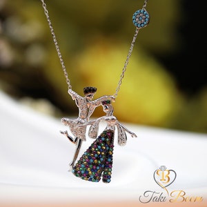 Può includere: Collana d'argento con un ciondolo che raffigura una coppia che balla. Il ciondolo è decorato con pietre preziose colorate. La collana ha anche un piccolo ciondolo di pietra blu.