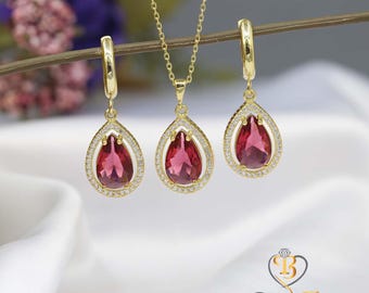 May include: A gold-toned jewelry set featuring a pendant necklace and matching earrings. Each piece showcases a teardrop-shaped, deep pink gemstone surrounded by a halo of small, clear stones. The set is displayed on a white satin surface.