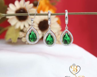 Puede incluir: Un conjunto de collar y pendientes de plata con piedras preciosas verdes en forma de lágrima. El colgante y los pendientes tienen un halo de pequeñas piedras transparentes. Las joyas se exhiben sobre un fondo rojo y blanco.