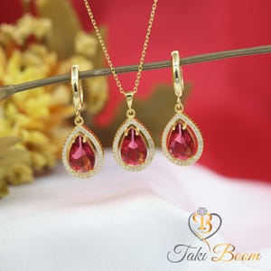 Puede incluir: Un conjunto de joyas doradas con un collar con colgante y pendientes a juego. Cada pieza presenta una piedra preciosa en forma de lágrima, de color rubí, rodeada por un halo de pequeños cristales transparentes. El conjunto se exhibe sobre un fondo rojo y blanco.