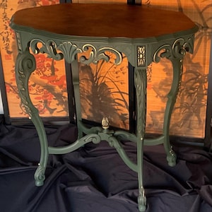 May include: A dark green side table with ornate gold accents and a wood top. The table has a unique, curved design and sits on a black surface.