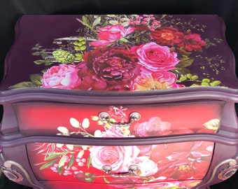 May include: A three-drawer dresser with a purple base and red drawers. The drawers are decorated with a floral pattern featuring pink and red roses.