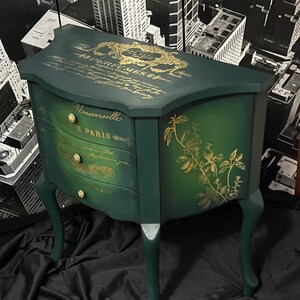 May include: A dark green, three-drawer cabinet with gold accents and floral designs. The cabinet has curved legs and a decorative top with gold script. The drawers have gold knobs and the text "Universelle DE PARIS".