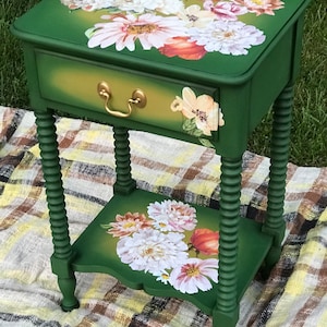 Op de afbeelding: Een groene bijzettafel met een lade en een onderplank. Zowel de lade als de plank hebben een bloemenmotief met witte, roze en oranje bloemen. De tafel heeft gedraaide poten.