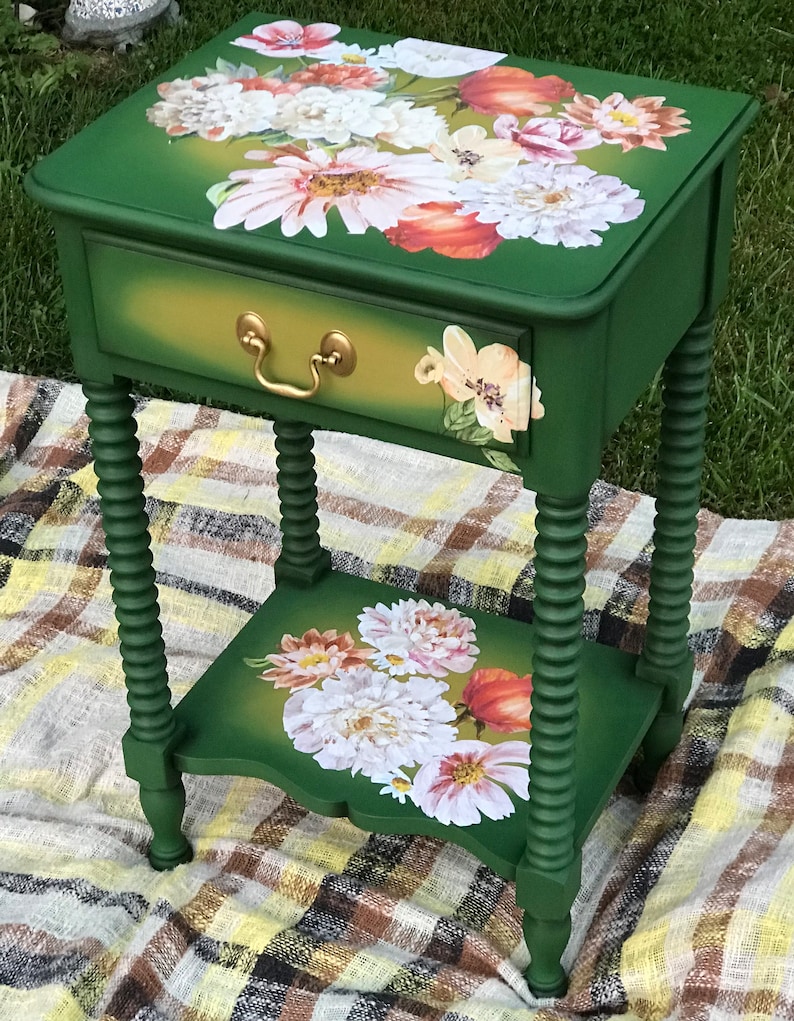 Op de afbeelding: Een groene bijzettafel met een lade en een onderplank. Zowel de lade als de plank hebben een bloemenmotief met witte, roze en oranje bloemen. De tafel heeft gedraaide poten.
