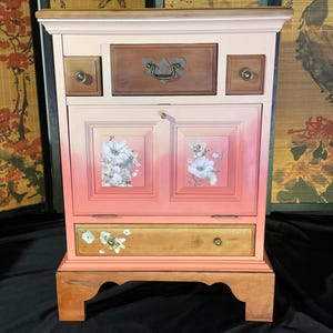 May include: A pink painted wooden cabinet with a floral design on the doors. The cabinet has three drawers, two small ones on the top and one larger one on the bottom. The cabinet is sitting on a wooden base.