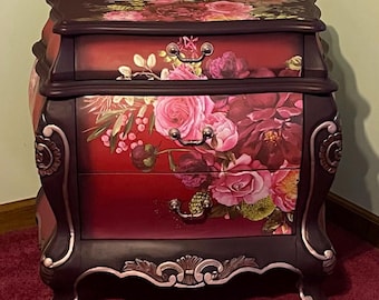 May include: A dark-toned wooden nightstand featuring three drawers with silver-colored metal pulls. The front and top surfaces are decorated with a vibrant floral print of pink and red roses against a deep red and black background.