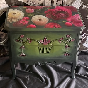 May include: A two-drawer green painted chest of drawers with floral decoupage on the top drawer and carved floral details on the front. The chest has a distressed finish and sits on four legs.