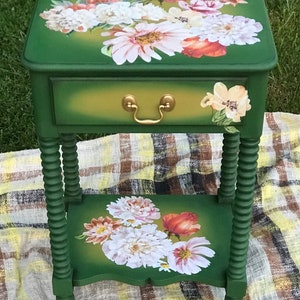 Op de afbeelding: Een groene bijzettafel met een lade en een onderplank. De tafel is versierd met een bloemenontwerp met witte, roze en oranje bloemen. De tafel heeft gedraaide poten.