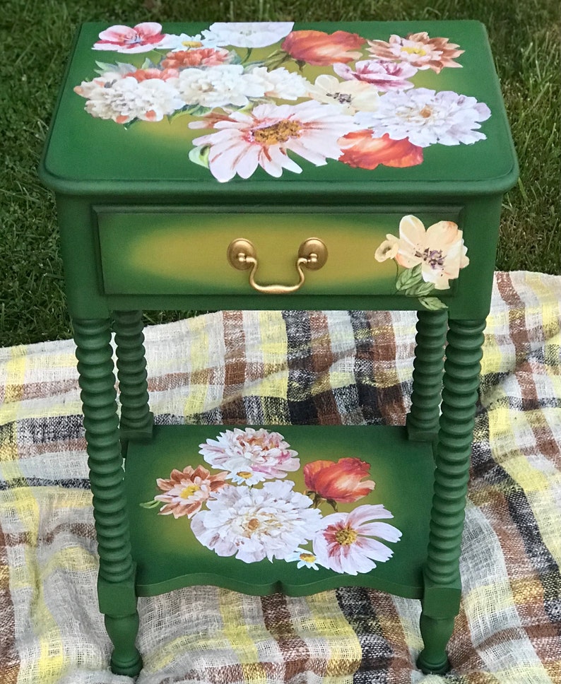 Op de afbeelding: Een groene bijzettafel met een lade en een onderplank. De tafel is versierd met een bloemenontwerp met witte, roze en oranje bloemen. De tafel heeft gedraaide poten.