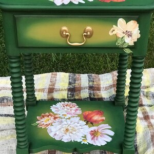 Op de afbeelding: Een groene bijzettafel met een lade en een onderplank. De tafel is versierd met bloemmotieven in tinten roze, wit en rood. De tafel heeft gedraaide poten en een gouden ladegreep.