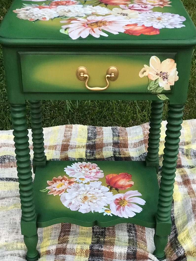 Op de afbeelding: Een groene bijzettafel met een lade en een onderplank. De tafel is versierd met bloemmotieven in tinten roze, wit en rood. De tafel heeft gedraaide poten en een gouden ladegreep.