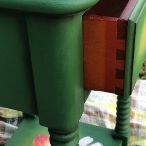 Op de afbeelding: Een groene bijzettafel met een lade en gedraaide poten. Het tafelblad heeft een bloemmotief met klaprozen en madeliefjes.