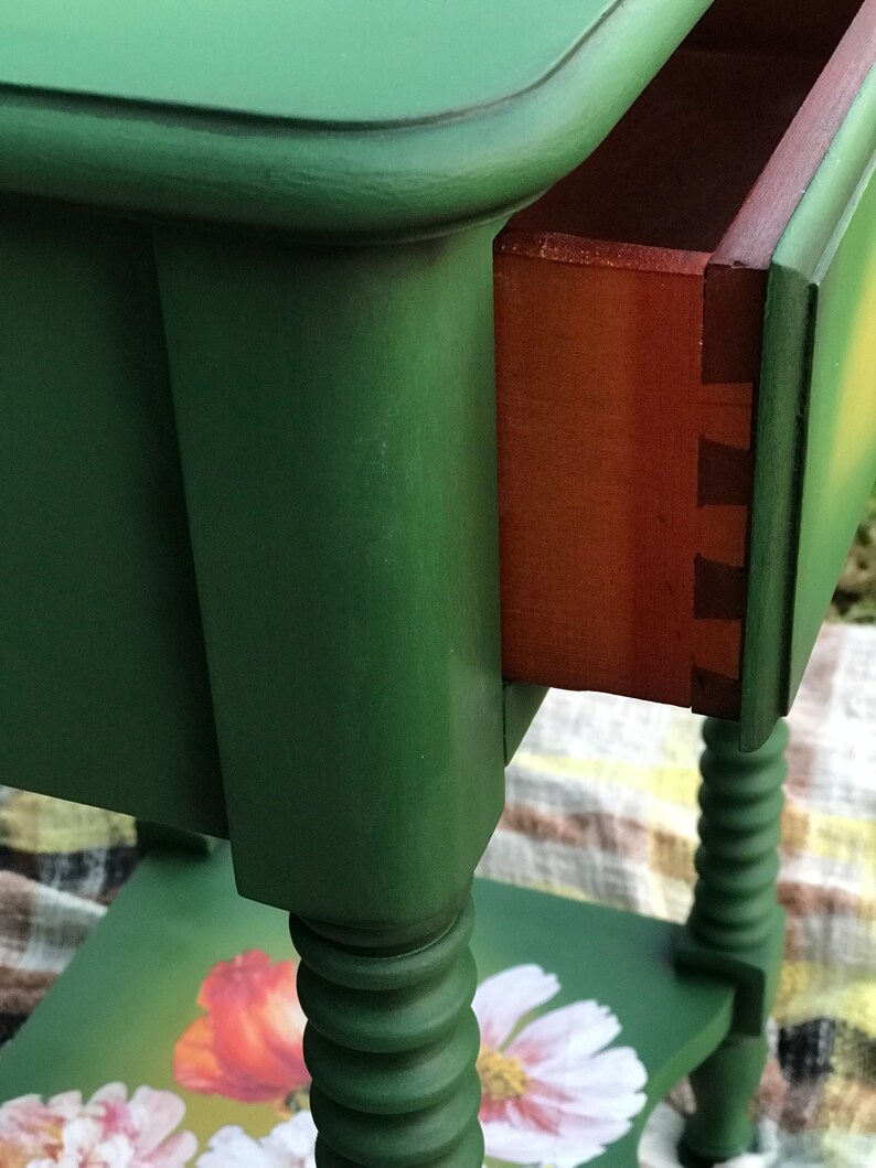 Op de afbeelding: Een groene bijzettafel met een lade en gedraaide poten. Het tafelblad heeft een bloemmotief met klaprozen en madeliefjes.
