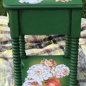 Op de afbeelding: Een groene bijzettafel met een bloemenontwerp op de bovenste en onderste plank. De tafel heeft gedraaide poten en een lade.
