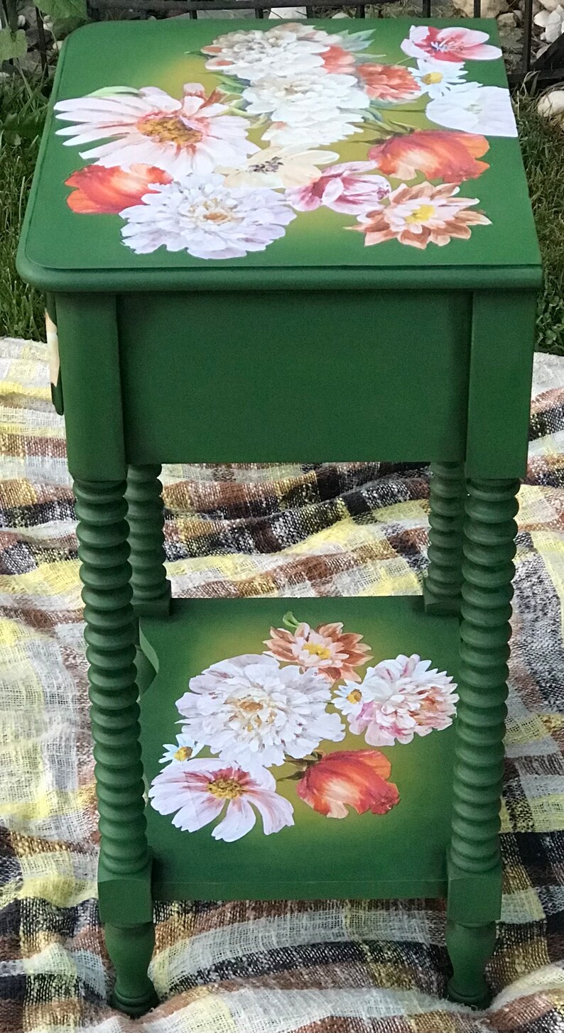 Op de afbeelding: Een groene bijzettafel met een bloemenontwerp op de bovenste en onderste plank. De tafel heeft gedraaide poten en een lade.