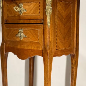 May include: A small, antique wooden nightstand with two drawers. The nightstand has a light brown finish with a decorative inlaid pattern. It features ornate brass hardware, including drawer pulls and corner accents. The legs are curved and slender, adding to its elegant design.