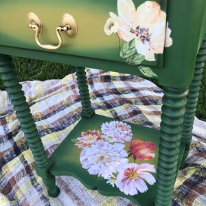 Op de afbeelding: Een groene bijzettafel met een lade en een onderplank. De lade heeft een gouden handvat en is versierd met een wit bloemenontwerp. De plank is ook versierd met een bloemenontwerp.