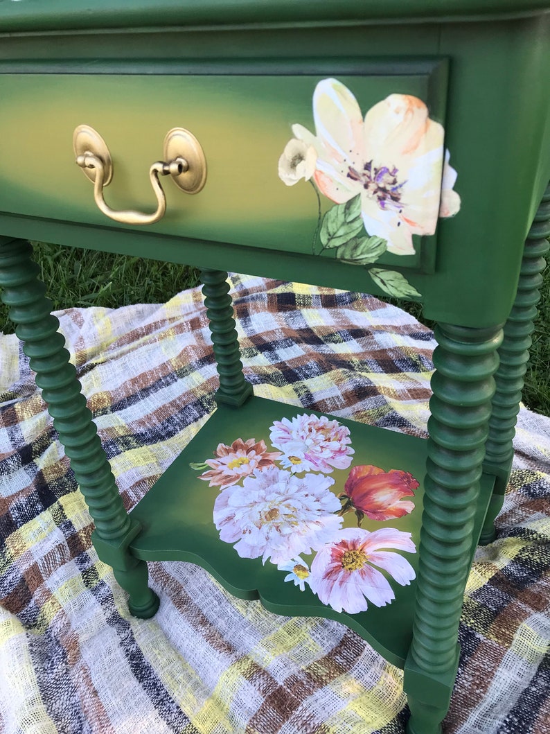 Op de afbeelding: Een groene bijzettafel met een lade en een onderplank. De lade heeft een gouden handvat en is versierd met een wit bloemenontwerp. De plank is ook versierd met een bloemenontwerp.