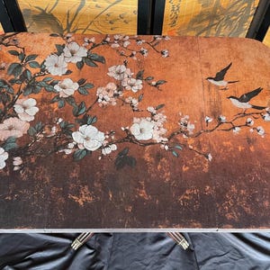 May include: A rectangular folding table with a floral and bird design. The table features a warm brown background with white blossoms and black birds. The table has silver legs and is set against a black background.