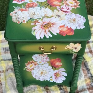 Op de afbeelding: Een groene bijzettafel met een lade en gedraaide poten. De bovenkant van de tafel is versierd met een bloemenontwerp met witte, roze en oranje bloemen.
