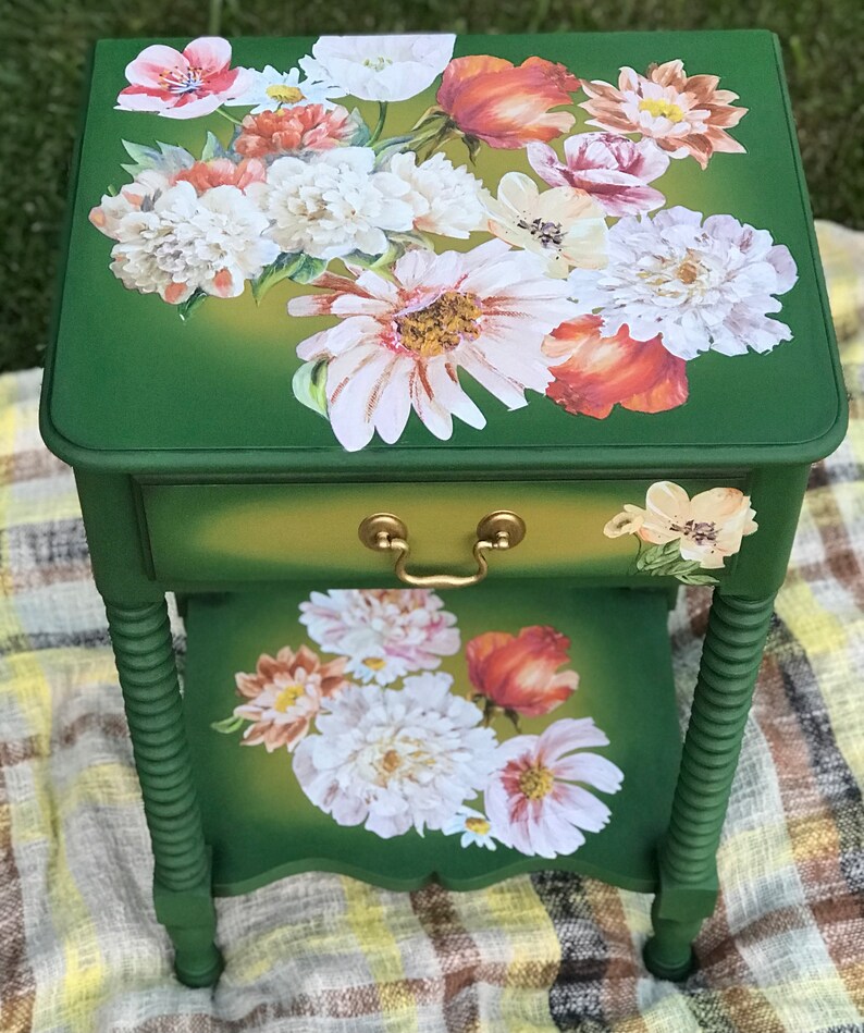Op de afbeelding: Een groene bijzettafel met een lade en gedraaide poten. De bovenkant van de tafel is versierd met een bloemenontwerp met witte, roze en oranje bloemen.