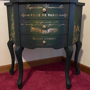 May include: A dark green three-drawer nightstand with curved legs and gold-colored lettering and floral designs. The drawers feature the text Exposition Universelle, VILLE DE PARIS, and Maison Clément, accented by small round metallic knobs.