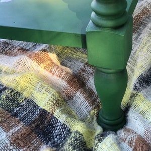 Op de afbeelding: Een groene geverfde houten tafel met een bloemenontwerp op de bovenkant. De tafel heeft een gedraaid poot en staat op een geruite deken.
