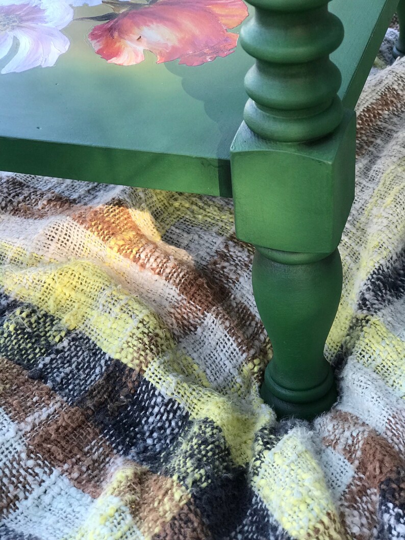 Op de afbeelding: Een groene geverfde houten tafel met een bloemenontwerp op de bovenkant. De tafel heeft een gedraaid poot en staat op een geruite deken.