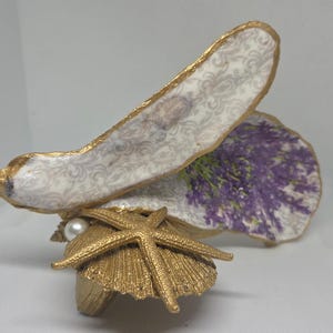 Puede incluir: Una escultura decorativa de concha de ostra con un borde pintado en oro y una superficie blanca con un patrón de damasco gris y un estampado floral de lavanda púrpura. La base está adornada con una estrella de mar dorada y una perla blanca.