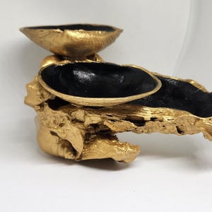 Puede incluir: Una pieza decorativa artesanal con una base texturizada dorada y tres cuencos negros y dorados. Los cuencos son de diferentes tamaños y formas, creando una exhibición visualmente interesante. La pieza probablemente está hecha de madera.