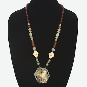 May include: A long necklace with a jade and bone pendant featuring a carved figure. The necklace is made of brown, green, and beige beads.