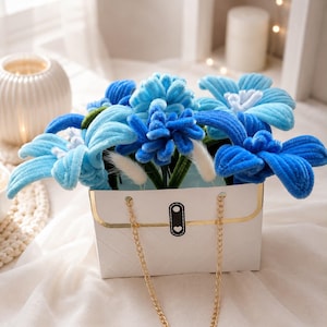 Op de afbeelding: Een witte gewatteerde handtas met een gouden ketting, gevuld met blauwe en lichtblauwe fluwelen bloemen. De bloemen hebben groene stelen en witte accenten, wat zorgt voor een levendig bloemstuk.